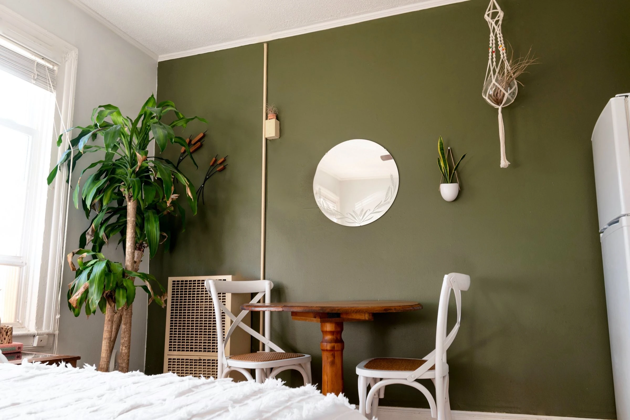 Un mur vert olive, un miroir rond, une table en bois et deux chaises blanches