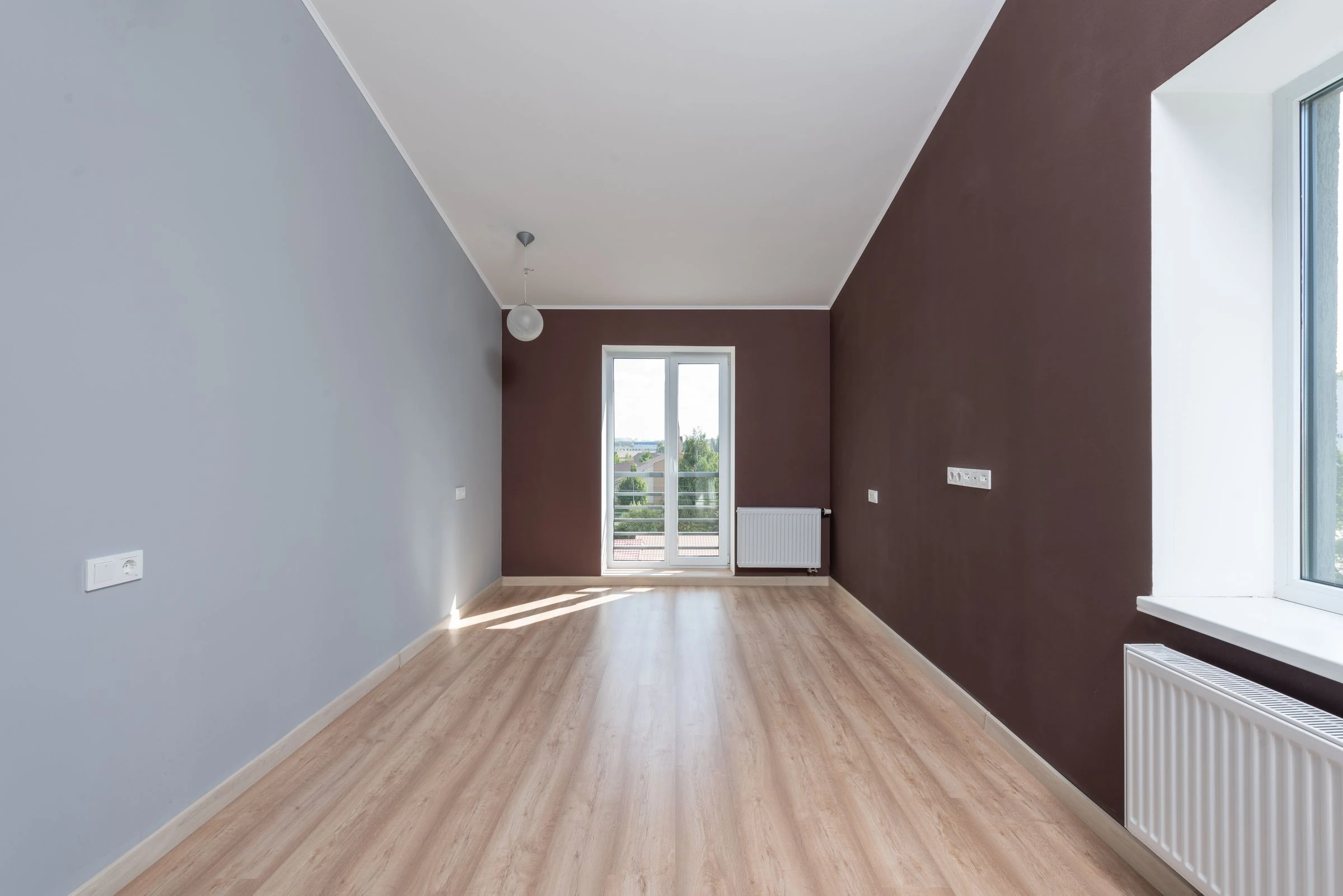 Une chambre vide aux murs gris bleu et brun, avec un parquet clair et une porte-fenêtre